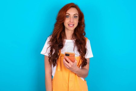 redhead woman wearing orange overall over blue background holds mobile phone in hands and rejoices positive news, uses modern cellular