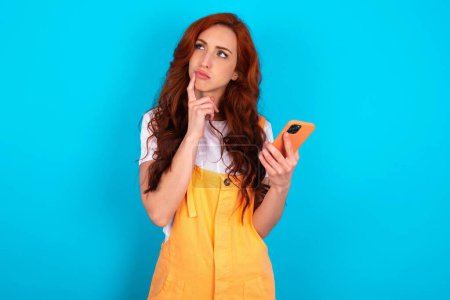 Téléchargez les photos : Rousse femme portant orange dans l'ensemble sur fond bleu pense profondément à quelque chose, utilise un téléphone mobile moderne, essaie de composer un bon message, garde l'index près des lèvres. - en image libre de droit