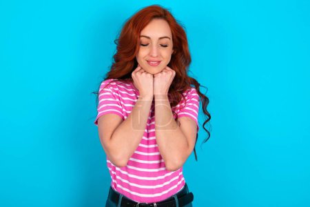 Photo for Cheerful redhead woman wearing pink striped T-shirt over blue background has shy satisfied expression, smiles broadly, shows white teeth, People emotions - Royalty Free Image