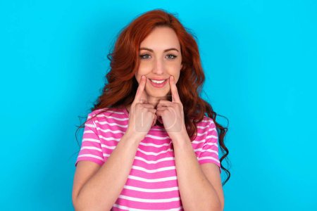 Foto de Mujer pelirroja feliz vistiendo camiseta de rayas rosadas sobre fondo azul con sonrisa dentada, mantiene los dedos índice cerca de la boca, los dedos señalando y forzando la sonrisa alegre - Imagen libre de derechos