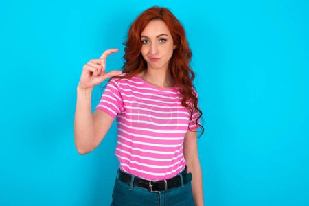 Téléchargez les photos : Femme rousse mécontente portant T-shirt à rayures roses sur fond bleu forme petit signe de la main démontre quelque chose de pas très grand. Concept de langage corporel. - en image libre de droit
