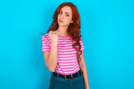 Photo for Redhead woman wearing pink striped T-shirt over blue background shows fist has annoyed face expression going to revenge or threaten someone makes serious look. I will show you who is boss - Royalty Free Image