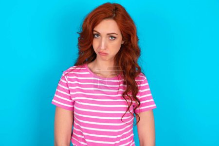 Foto de Pelirroja vistiendo una camiseta de rayas rosadas sobre fondo azul deprimida y preocupada por la angustia, llorando enojada y asustada. Expresión triste. - Imagen libre de derechos