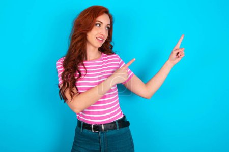 Foto de Pelirroja vistiendo camiseta de rayas rosadas sobre fondo azul sonrisa excitada dirigiendo los dedos mirada espacio vacío - Imagen libre de derechos