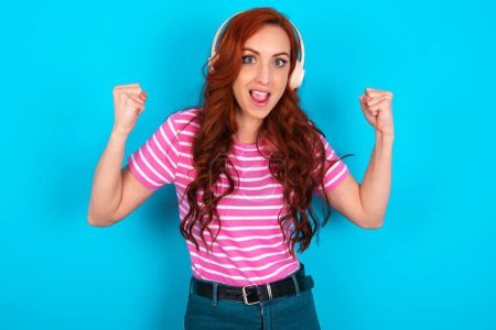 Photo for Emotional redhead woman wearing pink striped T-shirt over blue background exclaims loudly feels like winner raises clenched fists keeps mouth opened wears stereo headphones on ears makes yes gesture, listens favourite music - Royalty Free Image