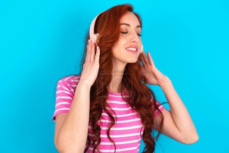 Foto de Pelirroja vistiendo una camiseta de rayas rosadas sobre fondo azul con auriculares en la cabeza, escucha música, disfruta de la canción favorita con los ojos cerrados, tomándose de la mano en los auriculares. - Imagen libre de derechos
