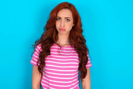 Photo for Displeased redhead woman wearing pink striped T-shirt over blue background frowns face feels unhappy has some problems. Negative emotions and feelings concept - Royalty Free Image