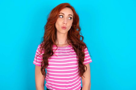 Foto de Mujer pelirroja impactada vistiendo camiseta de rayas rosadas sobre fondo azul mira espacio vacío con la boca abierta gritando: ¡Oh Dios mío! No puedo creerlo.. - Imagen libre de derechos