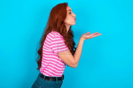Téléchargez les photos : Profil vue de côté portrait de jolie rousse femme portant rose rayé T-shirt sur fond bleu envoyer baiser d'air - en image libre de droit