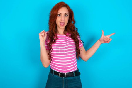 Foto de Pelirroja vistiendo camiseta de rayas rosadas sobre puntos de fondo azules en el espacio vacío sosteniendo el puño hacia arriba, gesto ganador. - Imagen libre de derechos