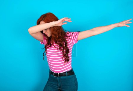 Photo for Photo of funky redhead woman wearing pink striped T-shirt over blue background show disco move dab - Royalty Free Image