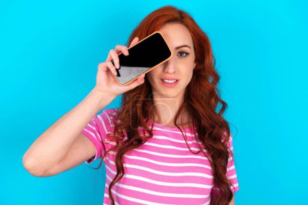 Foto de Pelirroja vistiendo una camiseta a rayas rosa sobre fondo azul sosteniendo un teléfono inteligente moderno cubriendo un ojo mientras sonríe - Imagen libre de derechos