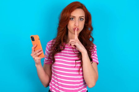 Photo for Redhead woman wearing pink striped T-shirt over blue background holding modern gadget ask not tell secrets - Royalty Free Image