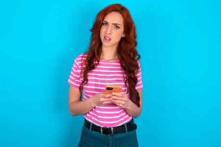 Téléchargez les photos : Photo de étonnante femme rousse folle portant T-shirt à rayures roses sur fond bleu tenir smartphone détestent concept de rétroaction - en image libre de droit