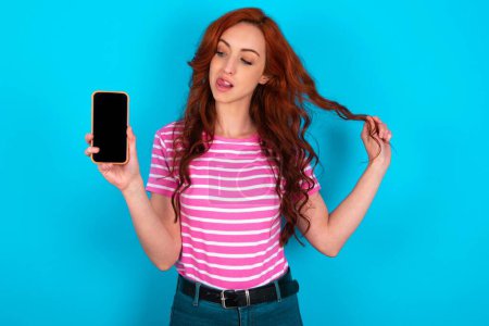 Photo for Photo of nice pretty redhead woman wearing pink striped T-shirt over blue background demonstrate phone screen hold hair tails - Royalty Free Image
