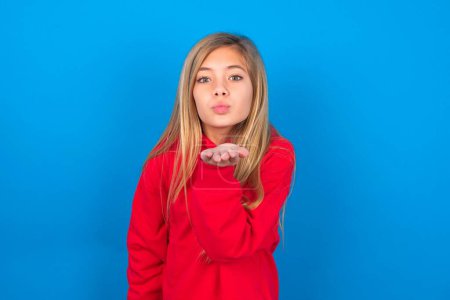 Téléchargez les photos : Belle adolescente caucasienne portant un sweat-shirt rouge sur fond bleu en regardant la caméra souffler un baiser avec la main sur l'air étant belle. Expression d'amour. - en image libre de droit