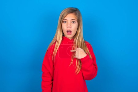 Shocked caucasian teen girl wearing red sweatshirt over blue background has astonished expression pointing at oneself with finger saying: Who me?