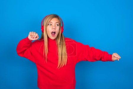 Foto de Bastante caucásico adolescente chica usando sudadera roja sobre fondo azul bailando y escuchando música con auriculares. - Imagen libre de derechos
