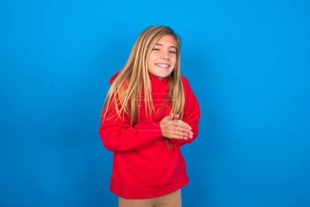 Foto de Chica adolescente caucásica con sudadera roja sobre fondo azul sintiéndose feliz, sonriendo y aplaudiendo, diciendo felicitaciones con un aplauso. - Imagen libre de derechos