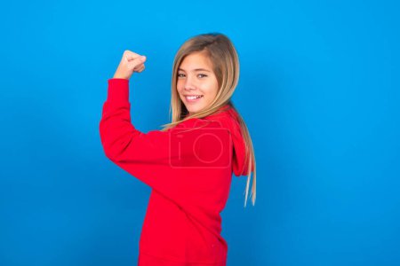 Foto de Encantadora chica adolescente caucásica con sudadera roja sobre fondo azul que muestra los músculos después del entrenamiento. Concepto de salud y fuerza. - Imagen libre de derechos