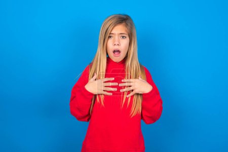 Caucasian teen girl wearing red sweatshirt over blue studio background keeps hands on chest feeling shocked and scared, mouth widely opened, stares at camera  