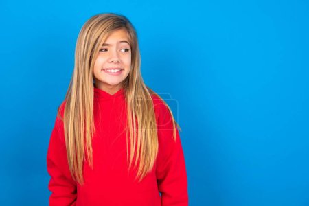 Foto de Retrato de niña adolescente caucásica con sudadera roja sobre fondo azul cierra los dientes y se ve confusamente a un lado, se da cuenta de su grave error - Imagen libre de derechos