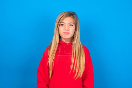 Téléchargez les photos : Caucasien adolescent fille portant rouge sweat-shirt sur fond bleu fait grimace avec maladroit expression a amusant seul, joue fou. - en image libre de droit