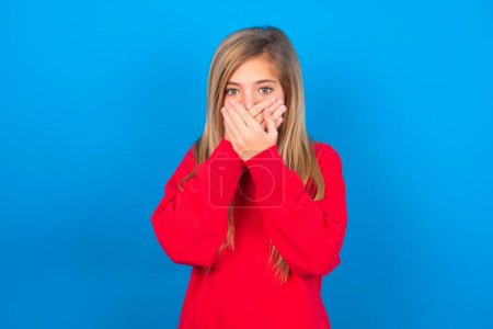 Téléchargez les photos : Bouleversé caucasien adolescent fille portant sweat-shirt rouge sur fond bleu couvrant sa bouche avec les deux paumes pour empêcher crier son, après avoir vu ou entendu quelque chose de mauvais. - en image libre de droit
