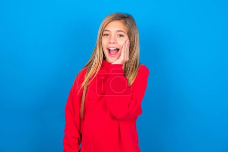 Photo for Shocked, astonished caucasian teen girl wearing red sweatshirt over blue background looking surprised in full disbelief wide open mouth with hand near face. Positive emotion facial expression body language. - Royalty Free Image