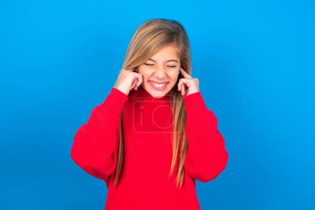 Téléchargez les photos : Malheureux stressé caucasien adolescent fille portant sweat-shirt rouge sur fond bleu faire face à l'inquiétude, bouchant les oreilles avec les doigts, irrité par un bruit fort. - en image libre de droit