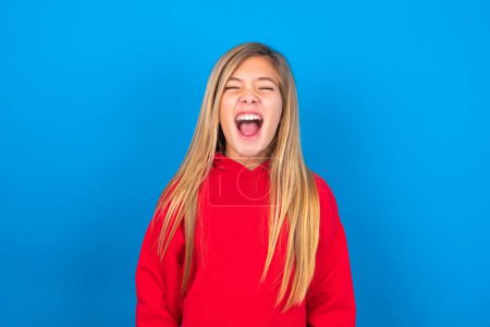 Photo for Stressful beautiful caucasian teen girl wearing red sweatshirt over blue background screams in panic, closes eyes in terror, finds out terrified news, can't believe it. - Royalty Free Image