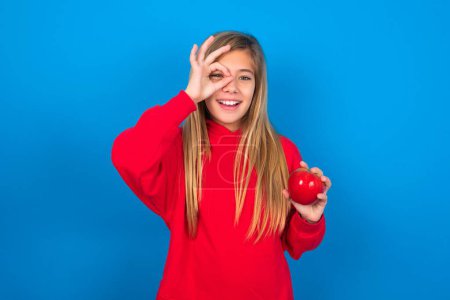 Foto de Hermosa chica adolescente caucásica con manzana usando sudadera roja sobre fondo azul con la cara feliz sonriendo haciendo signo ok con la mano en el ojo, mirando a través de los dedos. - Imagen libre de derechos