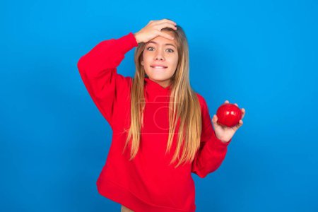 Photo for Blonde teen girl with apple wearing red sweatshirt over blue studio background holding hand on forehead with frightened and regret expression. - Royalty Free Image