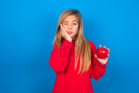 Foto de Rubia chica adolescente caucásica con sudadera roja sobre fondo azul con manzana en una mano y otra en la mejilla - Imagen libre de derechos