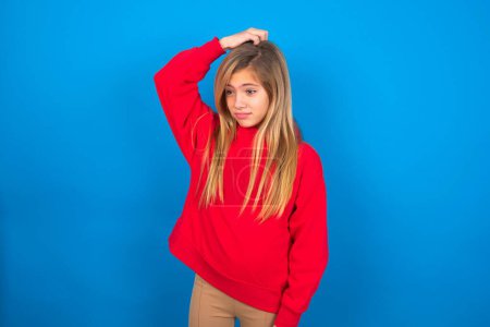 Téléchargez les photos : Belle adolescente caucasienne portant un sweat-shirt rouge sur fond bleu tenant la main sur la tête avec une expression de peur et de regret. - en image libre de droit