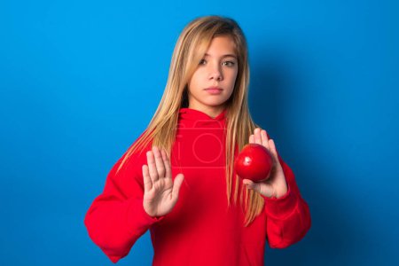 Photo for Beautiful caucasian teen girl wearing red sweatshirt over blue background holding an apple doing stop sing with palm of the hand. Warning expression with negative and serious gesture on the face. - Royalty Free Image
