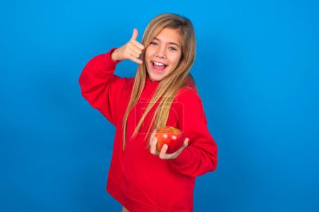 Photo for Beautiful caucasian teen girl wearing red sweatshirt over blue background holding an apple imitates telephone conversation, makes phone call gesture with hands, has confident expression. Call me! - Royalty Free Image