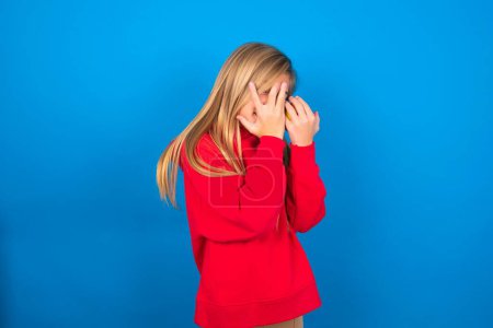 Photo for Pretty caucasian teen girl wearing red sweatshirt over blue background covering face with hands and peering out with one eye between fingers. Scared from something or someone. - Royalty Free Image