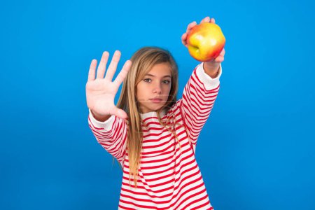 Foto de Retrato de una adolescente rubia sonriente con una camiseta a rayas sobre una pared azul mirando a la cámara y señalando el marco de los dedos. Concepto de creatividad y fotografía. - Imagen libre de derechos