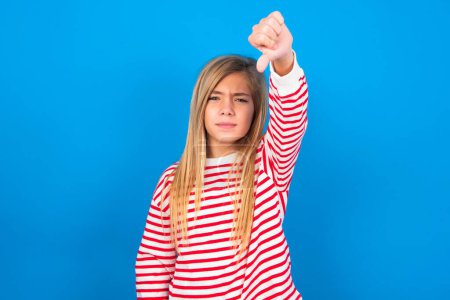 Photo for Discontent teen girl wearing striped shirt over blue background shows disapproval sign, keeps thumb down, expresses dislike, frowns face in discontent. Negative feelings. - Royalty Free Image