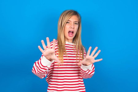 Photo for Dissatisfied teen girl wearing striped shirt over blue background frowns face, has disgusting expression, shows tongue, expresses non compliance, irritated with somebody. - Royalty Free Image