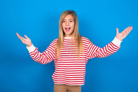 Téléchargez les photos : Joyeux adolescent fille portant une chemise rayée sur fond bleu faisant un geste de bienvenue levant les bras sur la tête. - en image libre de droit