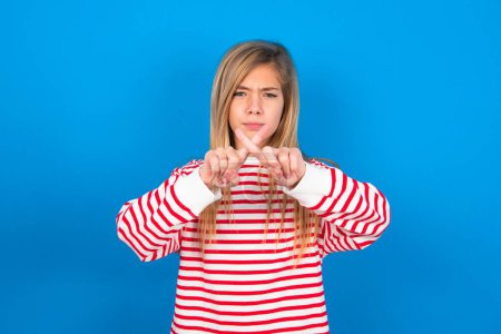 teen girl wearing striped shirt over blue background has rejection angry expression crossing fingers doing negative sign.