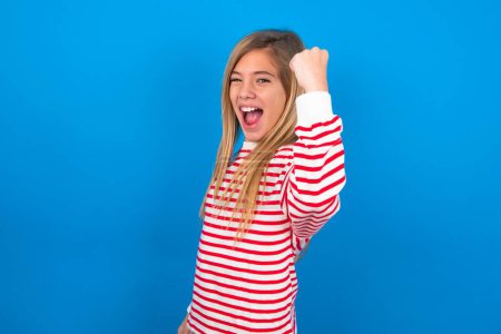 Photo for Overjoyed teen girl wearing striped shirt over blue background glad to receive good news, clenching fist and making winning gesture. - Royalty Free Image
