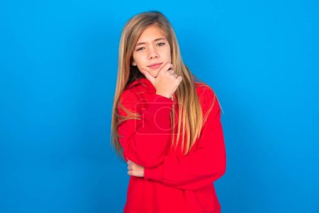Photo for Thoughtful caucasian teen girl wearing red sweatshirt over blue background keeps hand under chin, looks directly at camera, listens something with interest. Youth concept. - Royalty Free Image