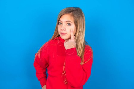 Téléchargez les photos : Adolescent caucasien fille portant sweat-shirt rouge sur fond bleu studio garder un oeil sur un objet devant, ou regarder dehors pour quelque chose - en image libre de droit