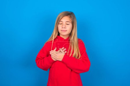 Photo for Beautiful caucasian teen girl wearing red sweatshirt over blue background closes eyes and keeps hands on chest near heart, expresses sincere emotions, being kind hearted and honest. Body language and real feelings concept. - Royalty Free Image