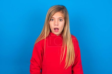 Photo for Caucasian teen girl wearing red sweatshirt over blue background having stunned and shocked look, with mouth open and jaw dropped exclaiming: Wow, I can't believe this. Surprise and shock - Royalty Free Image
