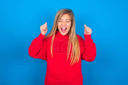 Téléchargez les photos : Belle adolescente caucasienne portant un sweat-shirt rouge sur fond bleu se réjouissant de son succès et de sa victoire serrant les poings avec joie étant heureuse d'atteindre l'objectif et les objectifs. Émotions positives, sentiments. - en image libre de droit