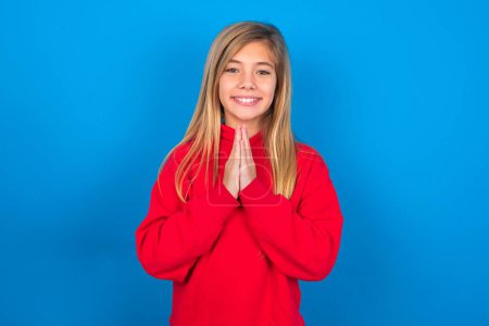 Photo for Charming caucasian teen girl wearing red sweatshirt over blue background keeps palms together, has pleased expression. Glad attractive male makes request, pleads for mercy. Hopeful young adult. - Royalty Free Image
